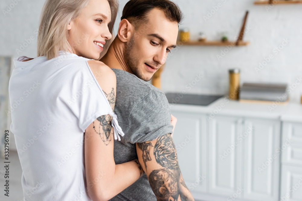Naklejka premium Back view of smiling woman embracing tattooed boyfriend in kitchen