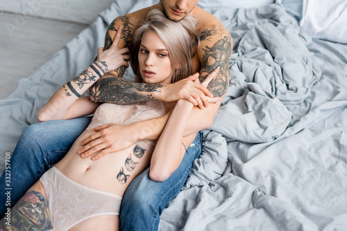 Muscular man in jeans embracing tattooed woman in underwear on bed