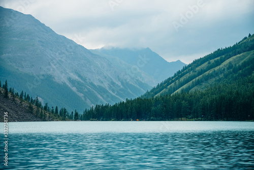 Dramatic view to vast mountain lake among giant mountains in rainy weather. Pines and larches on hillside near azure water. Overcast landscape with turquoise alpine lake. Atmospheric highland scenery.