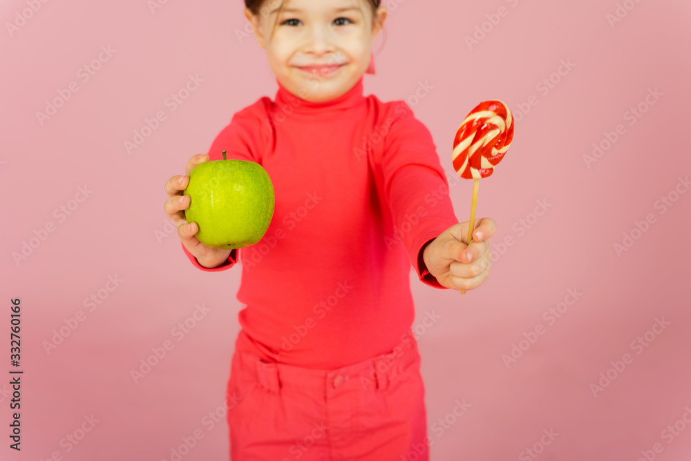 Little girl chooses between a lollipop and a green apple. The concept of proper nutrition. A child in a pink studio holds a sweet of sugar in his hand and an apple. Difficulty of choice Little girl chooses between a lollipop and a green apple. The concept of proper nutrition. A child in a pink studio holds a sweet of sugar in his hand and an apple. Difficulty of choice