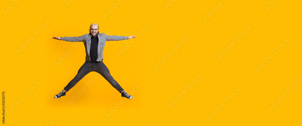 Attractive caucasian man with headphones and eyeglasses is jumping on a yellow wall with free space