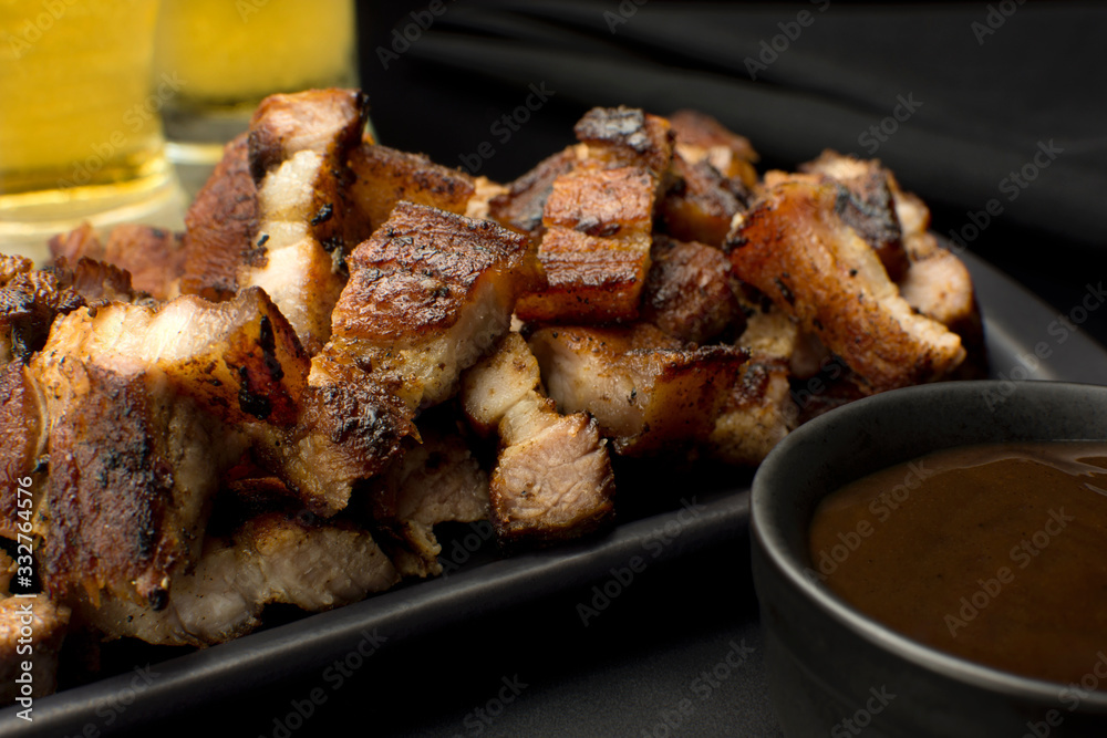 Grilled pork belly in a black platter with barbecue salce and beer in black background close
