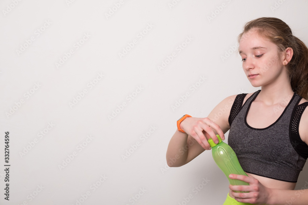 Positive young girl with a fitness bracelet drinks water from a bottle. Teen girl in sportswear holds a bottle of water. Sport girl in tank top for yoga drinks water after training. Positive young girl with a fitness bracelet drinks water from a bottle. Teen girl in sportswear holds a bottle of water. Sport girl in tank top for yoga drinks water after training.