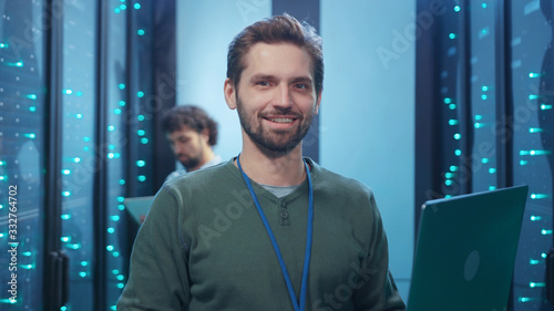 Fotografie Attractive cheerful young server specialist using laptop coworking in data center providing webhosting with colleague staying at computer security