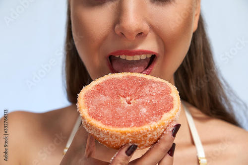Mouth of young woman bites grapefruit half in sugar in hand close up isolated on light background in studio