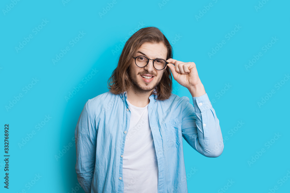 Long haired caucasian man with beard touching his glasses and looking at camera on a blue background Long haired caucasian man with beard touching his glasses and looking at camera on a blue background