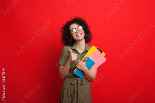 Curly haired caucasian student is holding some books and gesturing the like sign while posing on a red background