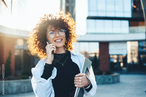 Curly haired caucasian businesswoman having a phone discussion while posing with eyeglasses and modern gadgets in front of a building photo