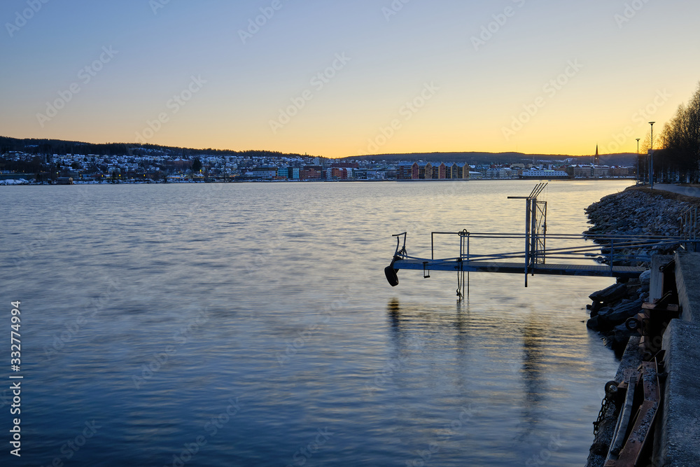 A small jetty goes out into the water with sunset and a city in the background