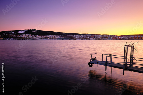 A small jetty goes out into the water with sunset and a city in the background