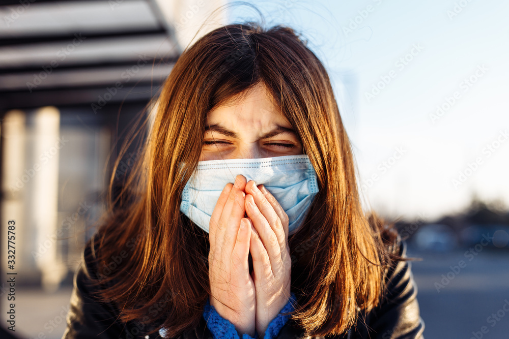 Young girl sneezes on a bus station and covers her face with hands. Girl wears a white medical mask standing near a bus at a public transport station. Coronavirus illness concept. Pandemia, epidemia. Young girl sneezes on a bus station and covers her face with hands. Girl wears a white medical mask standing near a bus at a public transport station. Coronavirus illness concept. Pandemia, epidemia.
