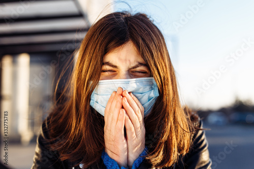 Young girl sneezes on a bus station and covers her face with hands. Girl wears a white medical mask standing near a bus at a public transport station. Coronavirus illness concept. Pandemia, epidemia.