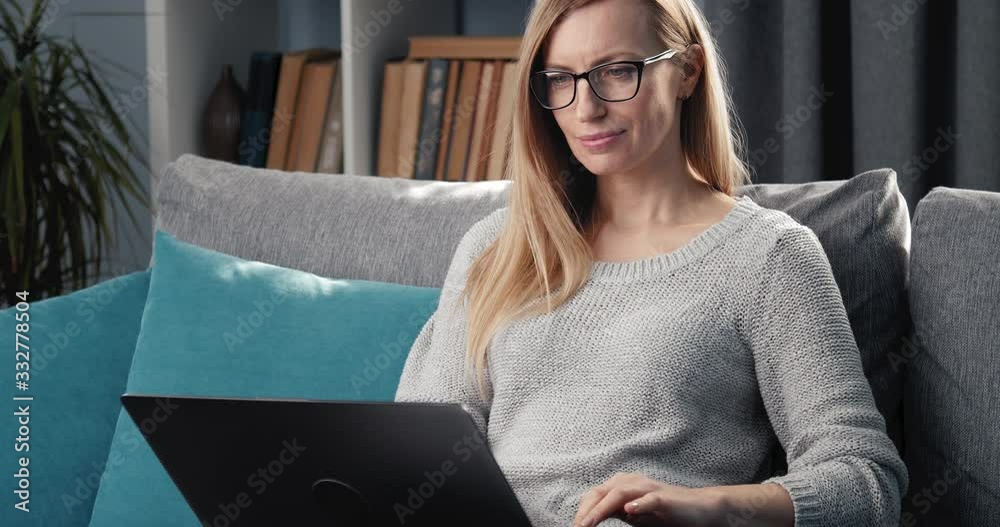 Mature business woman in casual clothing sitting on grey couch and working on laptop at home. Charming lady in eyeglasses solving urgent issues online.