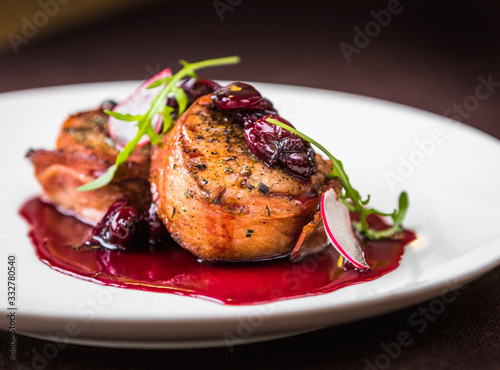 Pork medallions and bacon with cherry sauce on a white plate. Decorated with arugula and radish. Closeup