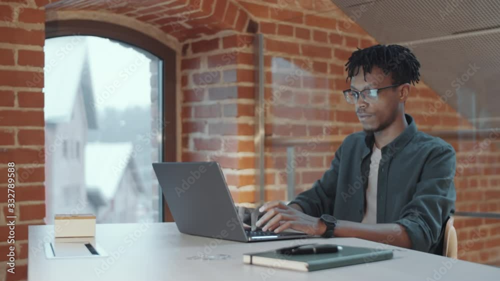 Cheerful young African American man with braids using laptop and then posing for camera with smile while working at desk near arch top window in loft office Cheerful young African American man with braids using laptop and then posing for camera with smile while working at desk near arch top window in loft office