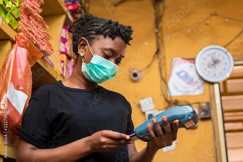 young african female attendant using the point of sale machine to pay for the goods her customer bought while using face mask to prevent herself from the corona outbreak.