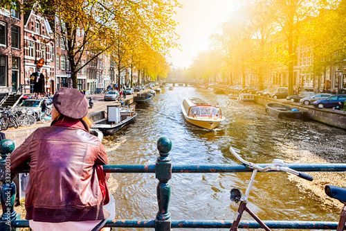 Traveling In Amsterdam City in the Netherlands. Back View of Female Sitting Near Amsterdam Canal Fence with a Torusit Boat in The Background photo