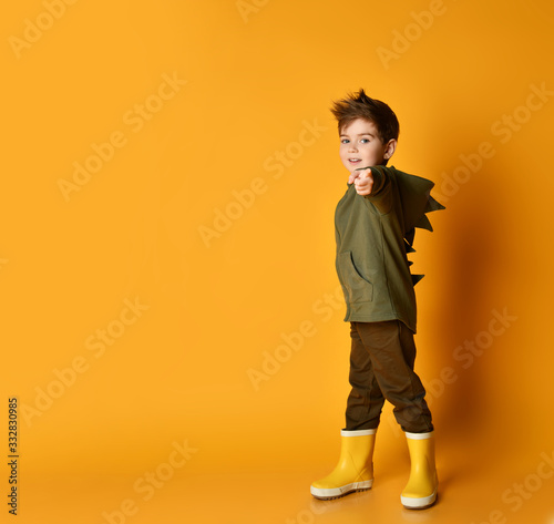 Cute kid boy in hoodie with dino spikes at back and cowl and in yellow rubber boots is pointing finger at us