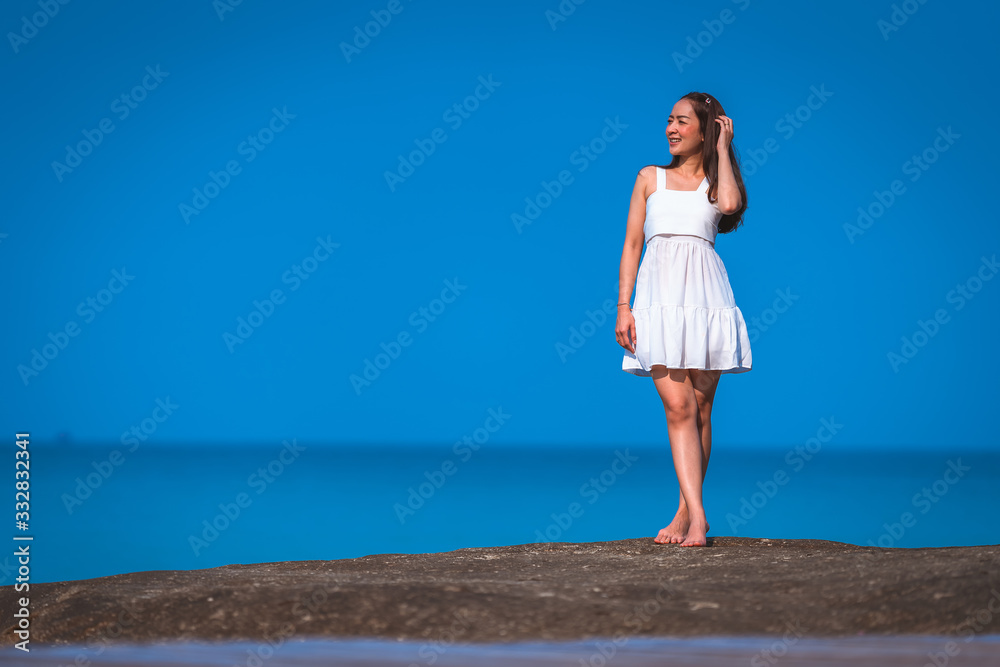 asian girl in white dress a holiday relax time in white dress by standing on stone on the sea with sunday light and blue sky in relaxing and freedom concept