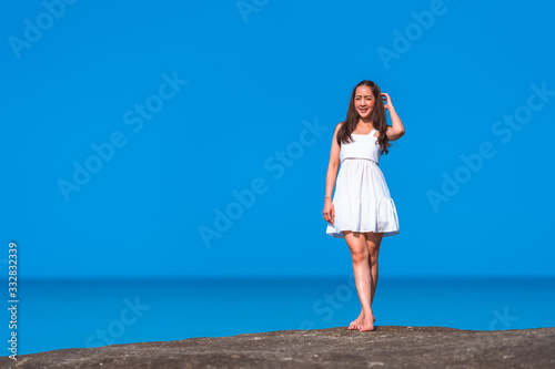 asian girl in white dress a holiday relax time in white dress by standing on stone on the sea with sunday light and blue sky in relaxing and freedom concept