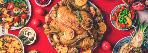 New year dinner table. Roasted Christmas chicken with orange slices, cranberries, garlic, festive decoration, candles, tangerine, pomegranate, golden glitter stars on red background. Top view, copy