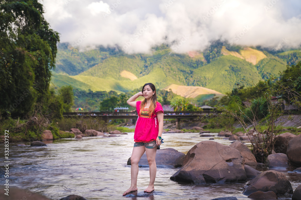 Naklejka premium Beautiful women wearing pink outfits standing on rocks in a stream surrounded by nature, with a background of mountains and clouds.