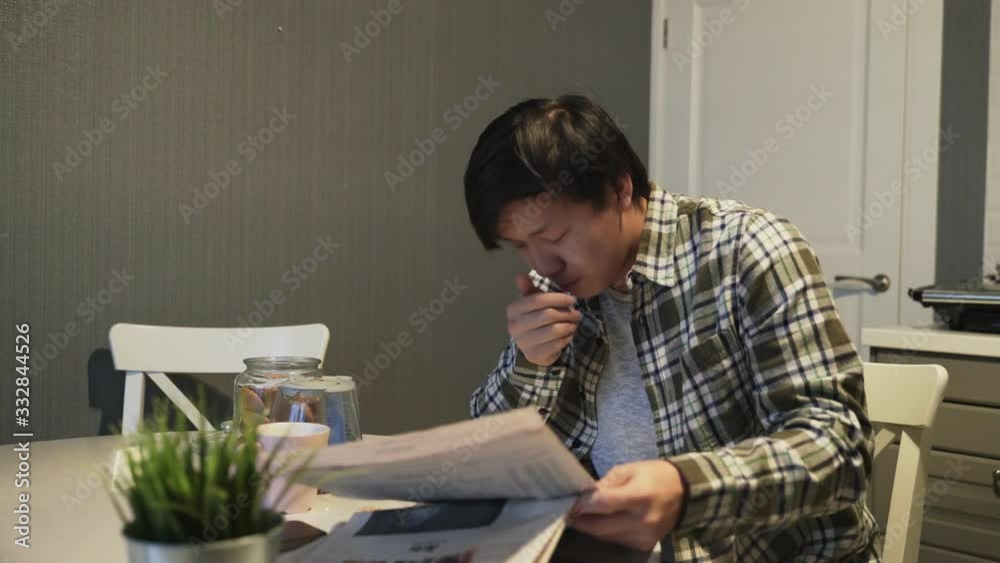 Sleepy asian man eating a cookie and reading newspaper at the table. Sleepy asian man eating a cookie and reading newspaper at the table.