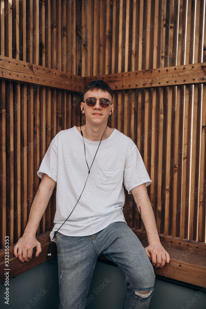 a young guy in dark glasses, a white t-shirt and blue jeans walks along the street and listens to music with headphones a young guy in dark glasses, a white t-shirt and blue jeans walks along the street and listens to music with headphones