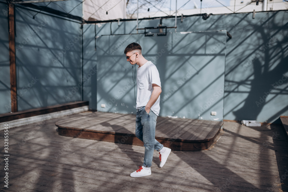 a young guy in dark glasses, a white t-shirt and blue jeans walks along the street and listens to music with headphones a young guy in dark glasses, a white t-shirt and blue jeans walks along the street and listens to music with headphones