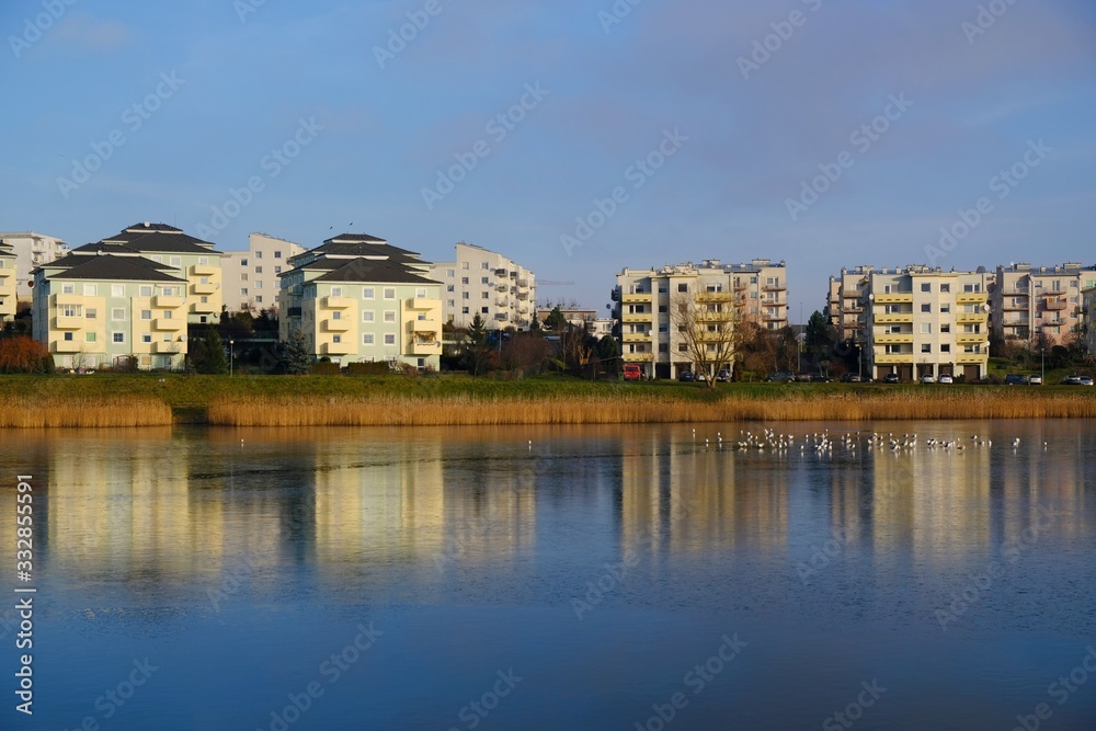 Fototapeta premium An estate of identical apartment blocks on the lake and its reflection in water, on the outskirts of the city. Idyllic view with blue sky and blue water. Gdansk South, Zakoniczyn, Poland