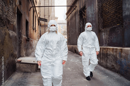 People with protective suits and respirators walking outdoors along deserted, abandoned, ruined street. Coronavirus concept.