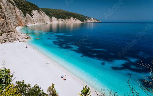 Summer vacation holiday. Fteri beach lagoon with rocky coastline, Kefalonia, Greece. Tourists under umbrellas chill relax near clear blue emerald turquoise sea lagoon