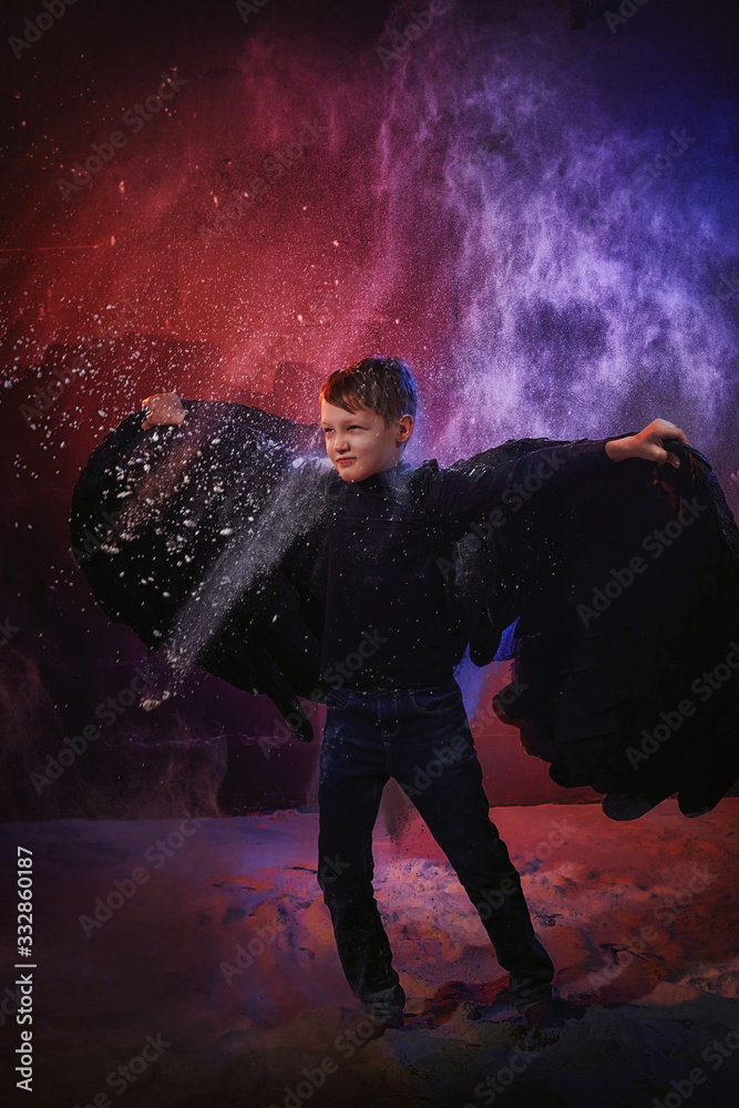 Black evil angel on a dark background with colored lighting. The concept of war between good and evil. Boy with angel wings during a photo shoot with flour and loose powder Black evil angel on a dark background with colored lighting. The concept of war between good and evil. Boy with angel wings during a photo shoot with flour and loose powder