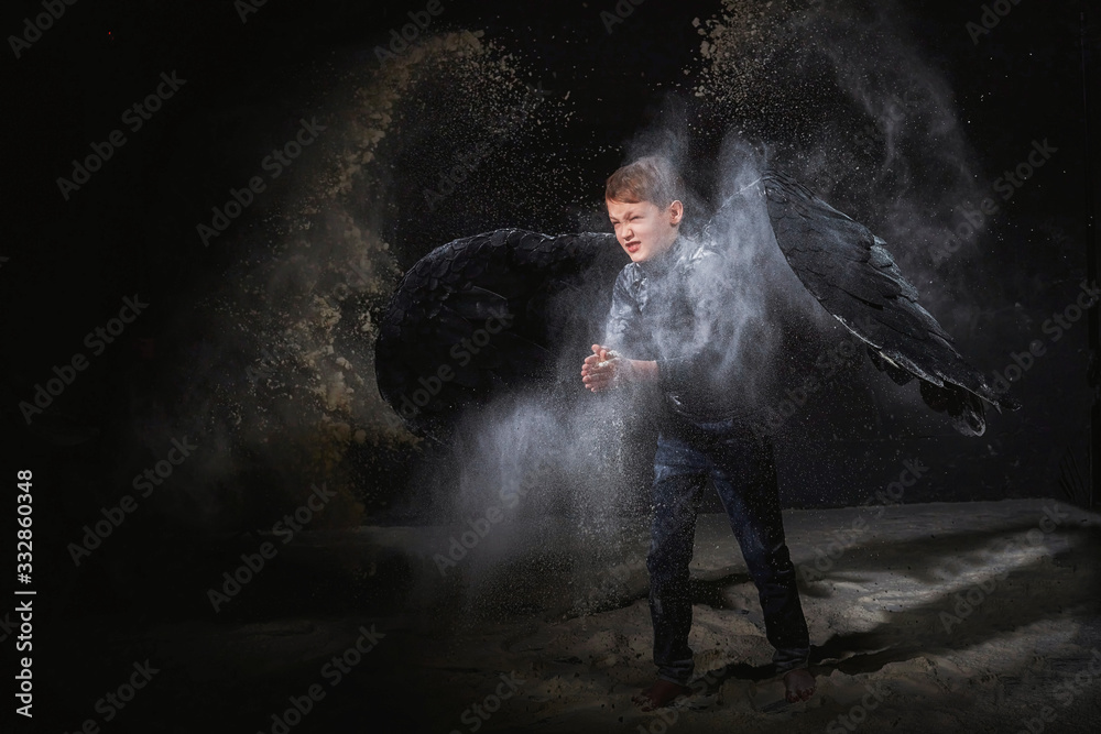 Black evil angel on a dark background with colored lighting. The concept of war between good and evil. Boy with angel wings during a photo shoot with flour and loose powder
