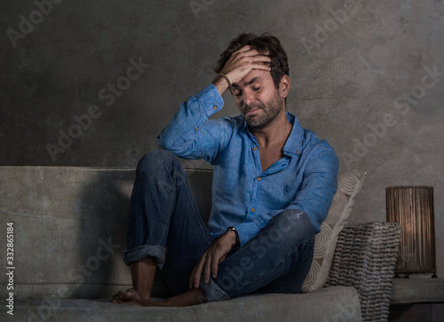 dark and dramatic lifestyle portrait of attractive depressed and worried man at home couch during covid-19 virus outbreak home lockdown feeling sad and overwhelmed