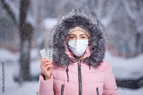 Portrait of a beautiful girl in a pink jacket in a medical protective mask in holding yellow pills in her hands for colds and flu. Winter street portrait of a woman under snowfall.