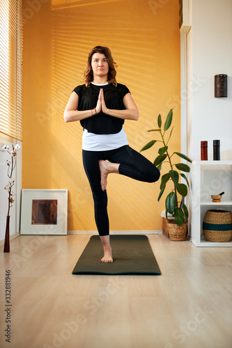 Middle aged brunette standing in Tree yoga pose at home in the morning.