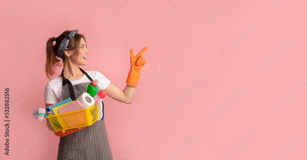 Cheerful Housewife Holding Basket With Cleaning Supplies And Pointing Aside Cheerful Housewife Holding Basket With Cleaning Supplies And Pointing Aside