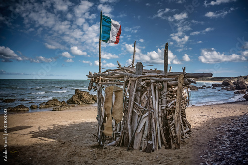 Cabane d'un Robinson Crusoé munie d'un drapeau français sur une plage. Illustration sur le confinement,l'isolement, leslogements précaires, sdf, sans-abris de France