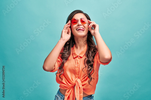 Beautiful young coquette woman dressed in orange shirt,red sunglasses with sticking tongue, looks at camera, wearing beige head wear. Cheerful and carefree concept.