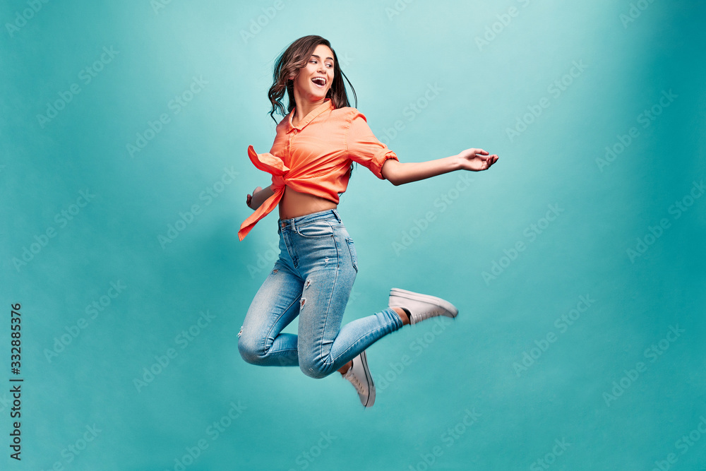 Catch fun emotions! Young beautiful woman who is dressed in an orange shirt and blue jeans bouncing on a blue background with happiness and looking away and laughing.
