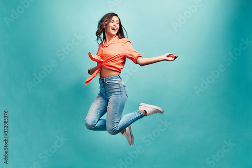 Catch fun emotions! Young beautiful woman who is dressed in an orange shirt and blue jeans bouncing on a blue background with happiness and looking away and laughing. © HBS
