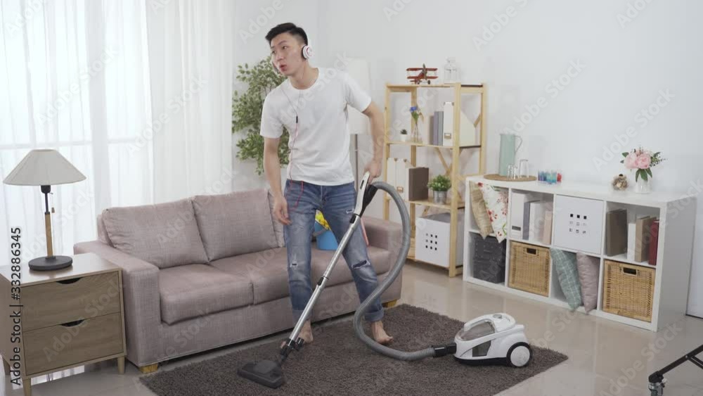 asian man with headphones is vacuuming living room and listening to online music streaming. chinese househusband cleaning house in a relaxed mood is humming and bobbing head with the upbeat song.