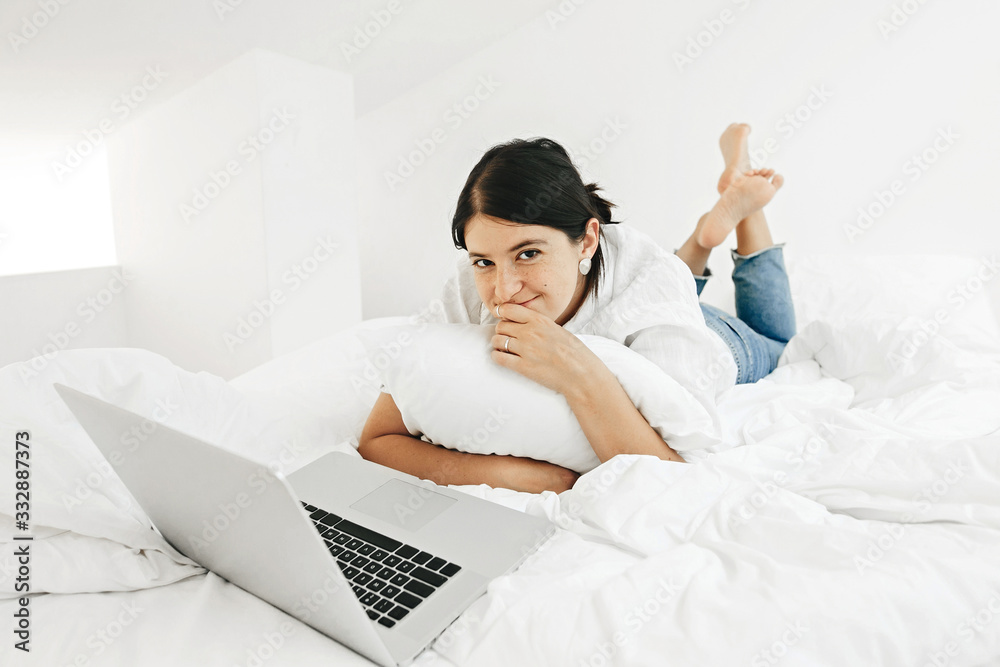 Happy stylish girl looking at laptop, lying on bed in white modern room. Young casual woman shopping online or studying online from home and thinking. Stay home. Freelance Happy stylish girl looking at laptop, lying on bed in white modern room. Young casual woman shopping online or studying online from home and thinking. Stay home. Freelance