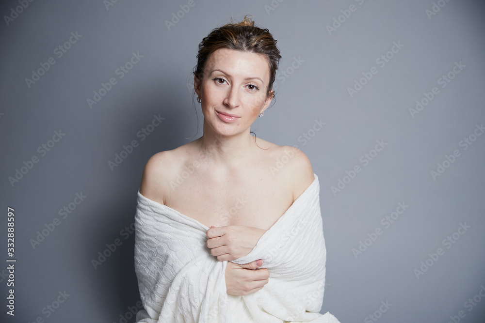 Portrait amazing slim cute beautiful young woman wrapped in white blanket with bare shoulders looks at camera on gray background Portrait amazing slim cute beautiful young woman wrapped in white blanket with bare shoulders looks at camera on gray background