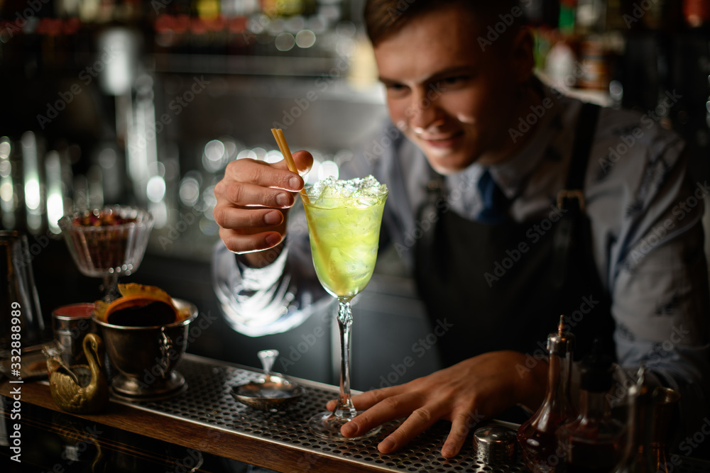 young smiling barman decorates by chopsticks wineglass with green cocktail.
