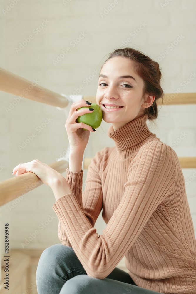 Young woman or pretty girl student, with cute young face and brown hair in casual outfit reading book, studying, eating apple near big window on sunny ballet dance room. Leisure and relaxing. Young woman or pretty girl student, with cute young face and brown hair in casual outfit reading book, studying, eating apple near big window on sunny ballet dance room. Leisure and relaxing.