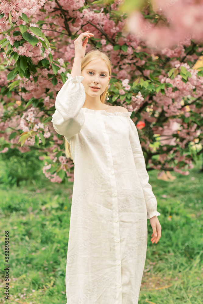 Gorgeous female model having fun in blooming garden. Natural background with blossom, warm sunlight, wellness and fitness concept. Sensual blond girl in white linen dress walking in Paris streets.