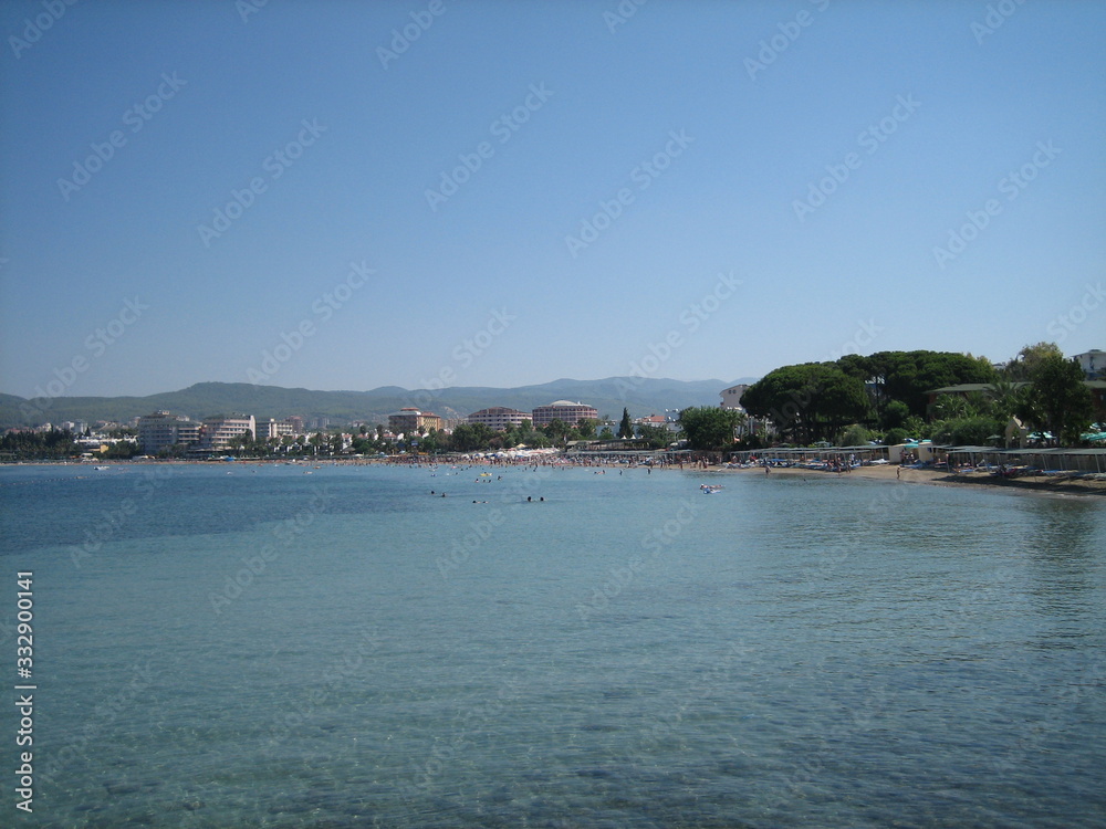 The coast of a calm sea with buildings and greenery on a cloudless day.