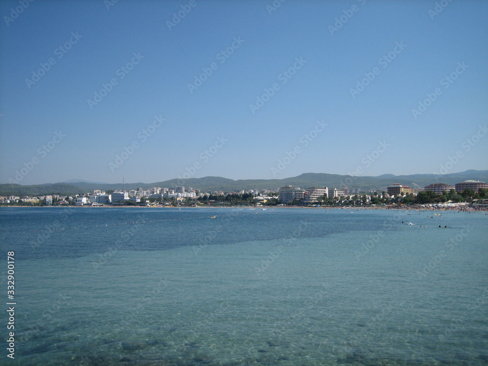 The coast of a calm sea with buildings and greenery on a cloudless day.